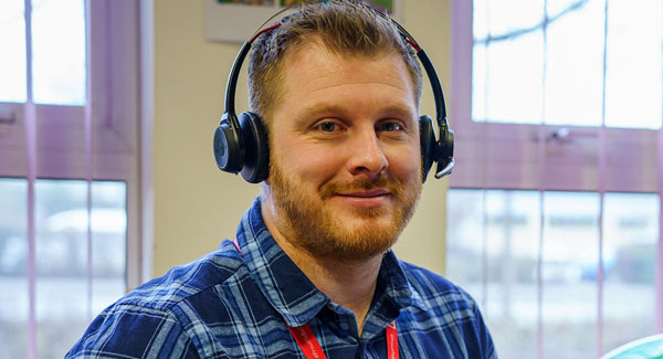 A man wearing a headset and blue checked shirt is sitting in the office smiling