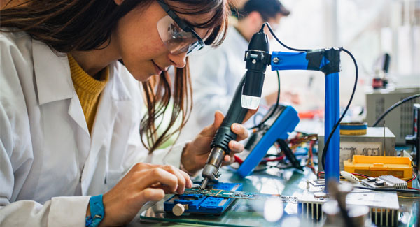A young female wearing safety goggles and a white lab coat is working on an electronic circuit board