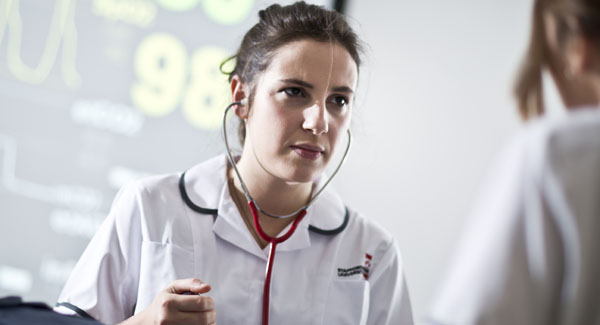 A female wearing a white nurse uniform and stethoscope is in a clinical setting