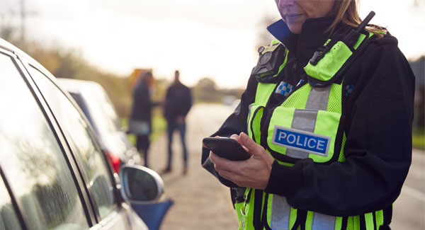 A female police officer wearing uniform is outside standing next to a car typing on a mobile phone