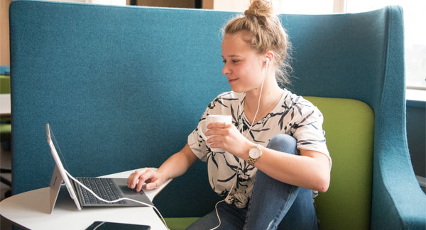 A female student with blonde hair sitting on a blue chair using a laptop and wearing earphones