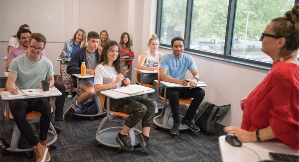A class of students wearing casual clothes sitting at individual desks in a lecture classroom with a female lecturer teaching at the front