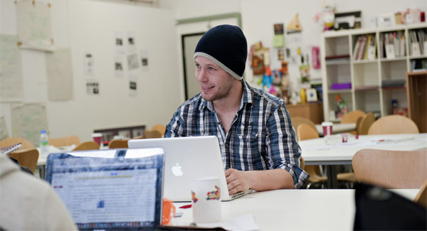 A male student wearing a hat and casual clothing sitting in a creative classroom with a laptop
