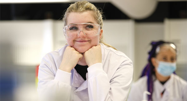 A female student with blonde hair is in the science lab wearing safety goggles and a white lab jacket