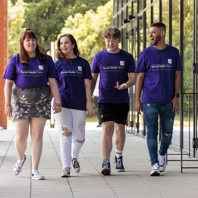 The Student Content Creator team composing of two females and two males wearing uniform purple t-shirts walking along the front of the Catalyst talking to each other on a bright day