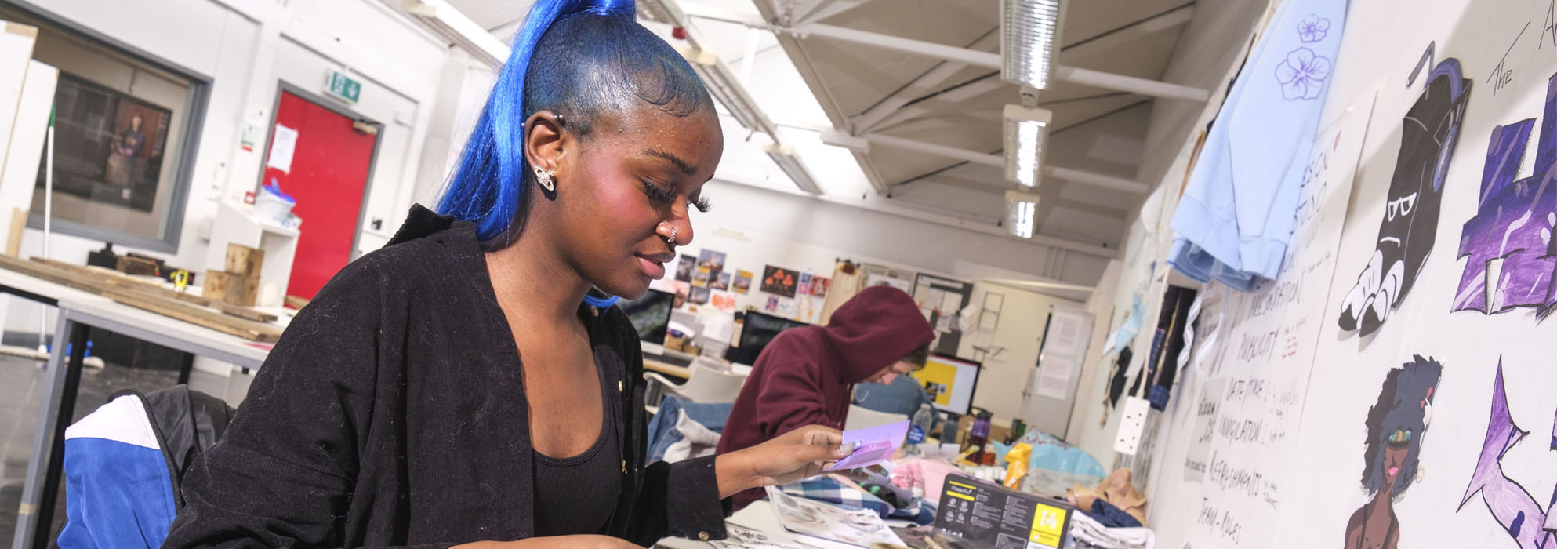 A female student with blue hair is working on a portfolio of printed graphics in the art and design workshop