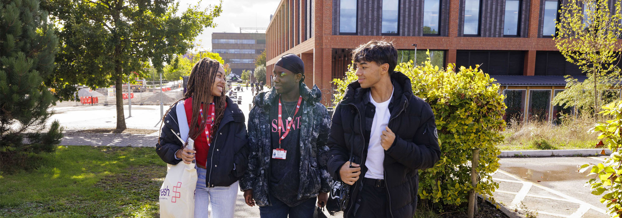 A group of three students are dressed casually and are talking and walking on the Stoke-on-Trent campus with the Catalyst building behind them