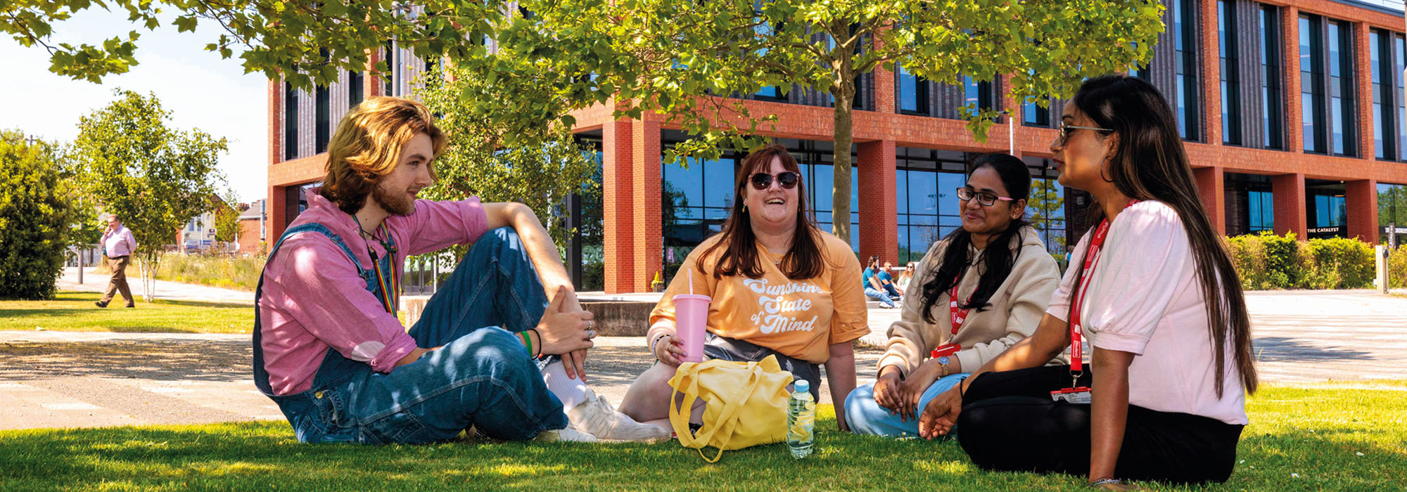 A group of four students wearing casual clothing sitting on a patch of grass talking to each other outside the Catalyst building on a bright day
