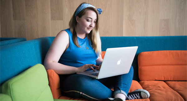 A female wearing a blue outfit and sitting on a colourful sofa is using a laptop to study