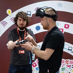 Male explains VR controls to another man who is wearing VR headset