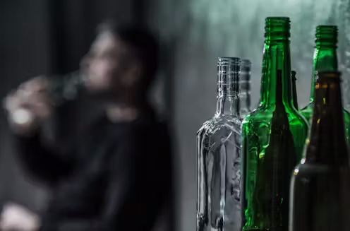 An image of alcohol bottles with a blurred figure drinking in the background