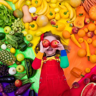 A young girl is surrounded by colourful fruit and vegetables and is holding 2 red apples