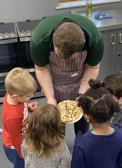 Young children are in the kitchen with a teacher, they are looking at a pancake on a plate