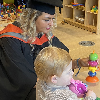 A female student with blonde hair is wearing graduation attire, playing in the nursery with her toddler
