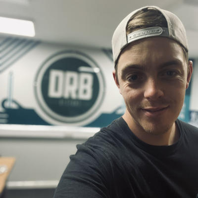 Daniel is smiling into the camera whilst wearing a dark blue t-shirt and a cream baseball cap backwards. Behind him is a blurred background with the letters "DRB" in large white writing within a dark blue circle.