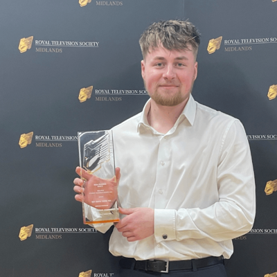 Duncan is smiling into the camera whilst wearing a white button shirt with long sleeves. Holding the award in his right hand, his left arm across his torso to hold the bottom of the award. The award is rectangular, glass in appearance, and with the Royal Television Society logo embossed at the top of the award. Behind Duncan is the golden RTS logo, with the words "Royal Television Society, Midlands" to the right of the logo. The imagery repeated multiple times across the backdrop.