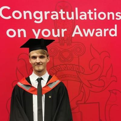 Fergus is wearing a cap and gown for graduation, smiling brightly into the camera. Behind him are the words "Congratulations on your Award" in white font on a red background.