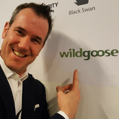 Johnn is wearing a white shirt and black suit, smiling brightly whilst pointing to the words "wildgoose" on a board behind him.