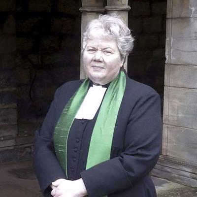 Reverend Elspeth McKay is standing in front of a stone building in traditional reverend attire, including a green stole. She is smiling into the camera with her hands held in front of her.