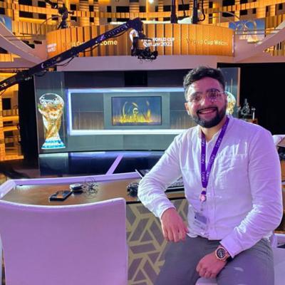 Saqib Uddin is sitting at a desk within a TV studio smiling brightly into a camera. Behind him is signage for the “World Cup Qatar 2022”.