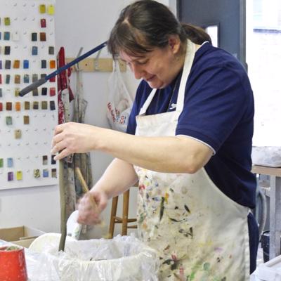 Sian is standing over a large bowl filled with clay. They are wearing a blue polo shirt which is covered by a white apron with various colours of paint on it. Behind Sian is an art studio with colour samples on the wall. 