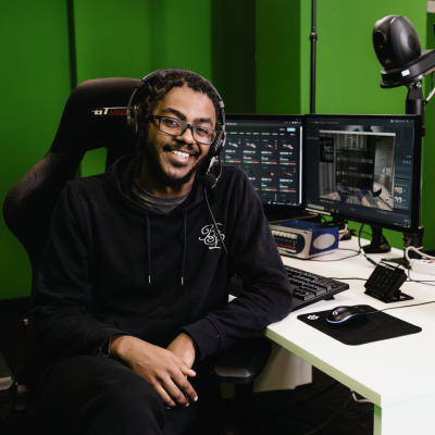 Andrew is wearing a black hoodie and sitting in a gaming chair. On the white desk are multiple monitors displaying CS:GO, along with a keyboard and mouse on the desk. Behind him is a green wall.