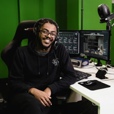 Andrew is wearing a black hoodie and sitting in a gaming chair. On the white desk are multiple monitors displaying CS:GO, along with a keyboard and mouse on the desk. Behind him is a green wall.
