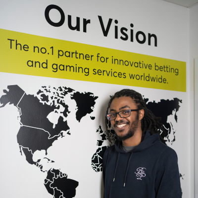 Andrew is wearing a black hoodie and standing in front of a white wall. On the wall in black are the words "Our Vision" at the top, then below is a yellow box with the words "The no.1 partner for innovative betting and gaming services worldwide.", then at the bottom is a large black map of the world.