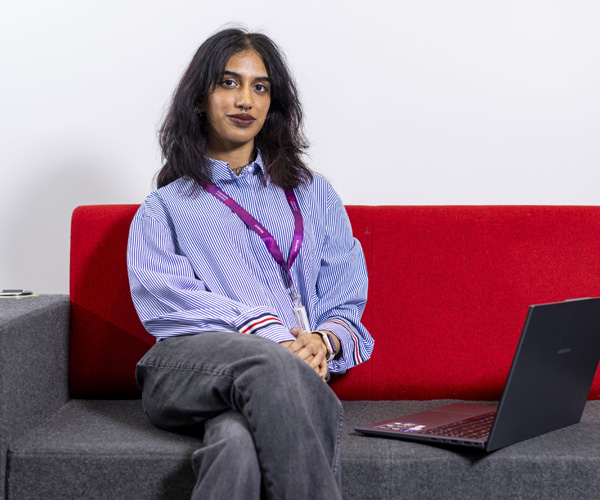 A female student with dark hair wearing a blue shirt is sitting on an office sofa with her laptop