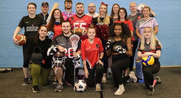 Students' Union representatives from across a variety of sports clubs are wearing their respective sports clothing and are in a hall together posing for a group photograph