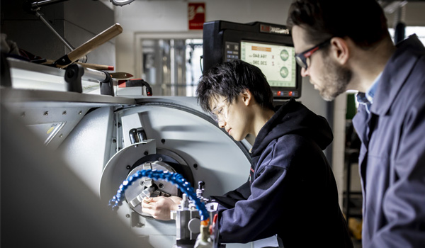 A male student wearing blue overalls and safety goggles is using engineering equipment in the lab
