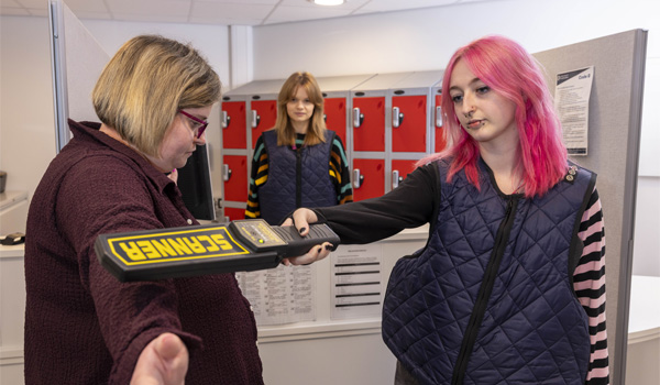 A female with pink hair is scanning a female with a detector in a security simulation
