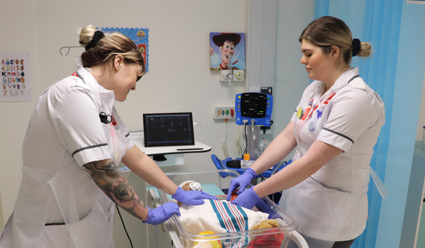 Two female nurses wearing white uniforms are caring for a dummy child in a cot