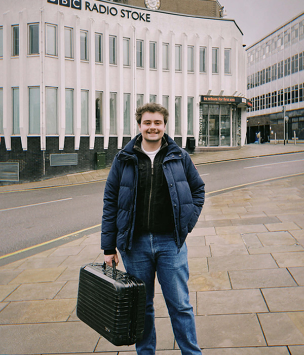 Brad is smiling and standing outside of BBC radio Stoke, he is holding a black briefcase
