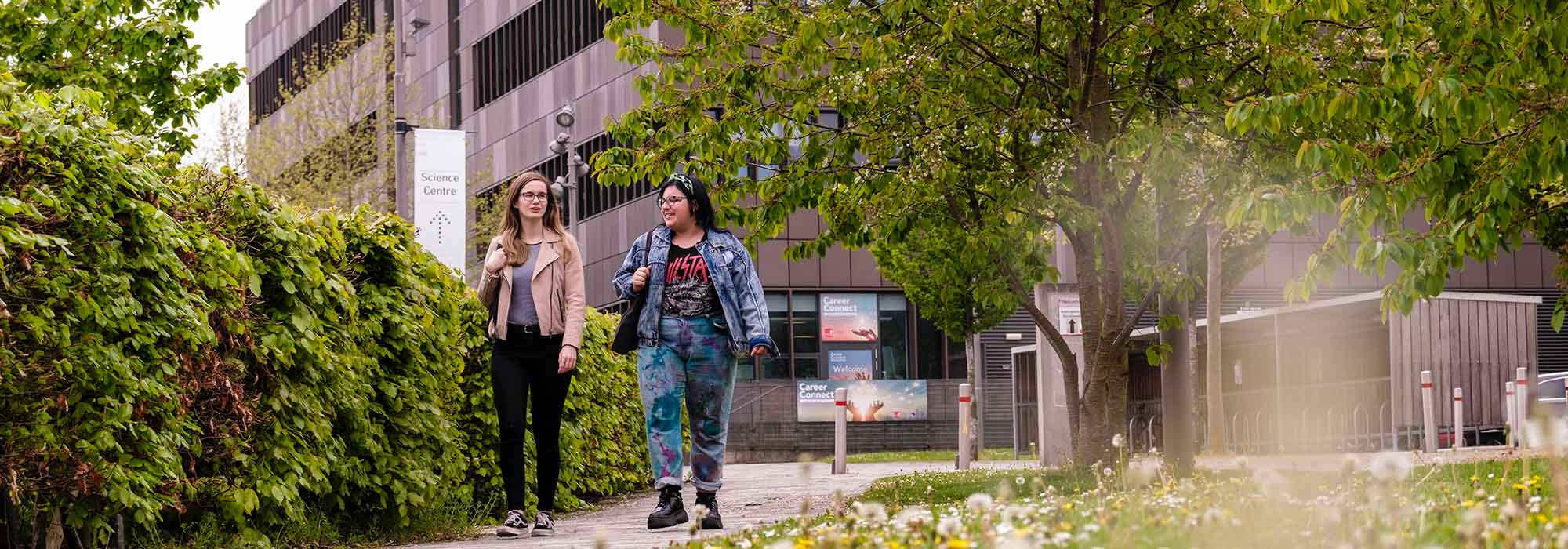 Two female students walking through our Leek Road site, in the background you can see the Science Centre and greenery