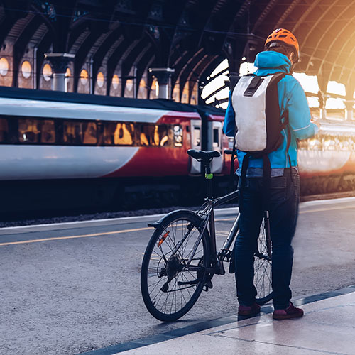 Male cyclist standing on a train station platform. He has a backpack and a brightly coloured helmet