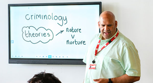 A university lecturer wearing a light coloured shirt standing at the front of class presenting a criminology lecture