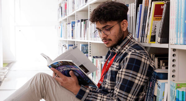 A male student wearing a blue and white checked shirt is sitting on the floor reading a book in front of the book shelves in the library