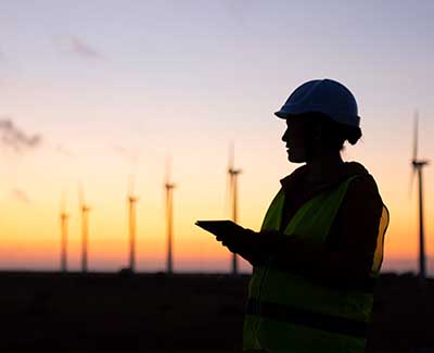 Female environmental engineer standing in front of a wind farm. She is wearing a hard hat and high visibility jacket and holds a clipboard.