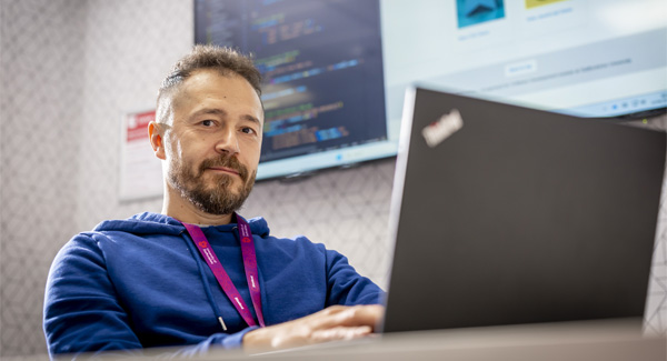 A male student with a beard wearing a blue shirt is using his laptop in a university room