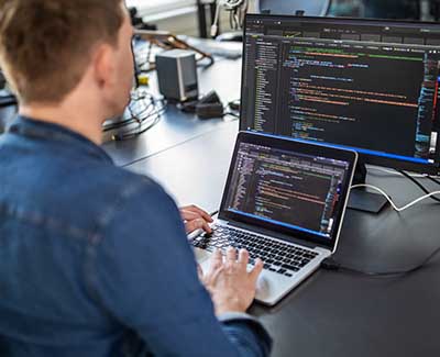 Young male programmer writes code on laptop, with a second monitor also showing code writing software. He is wearing a denim shirt.