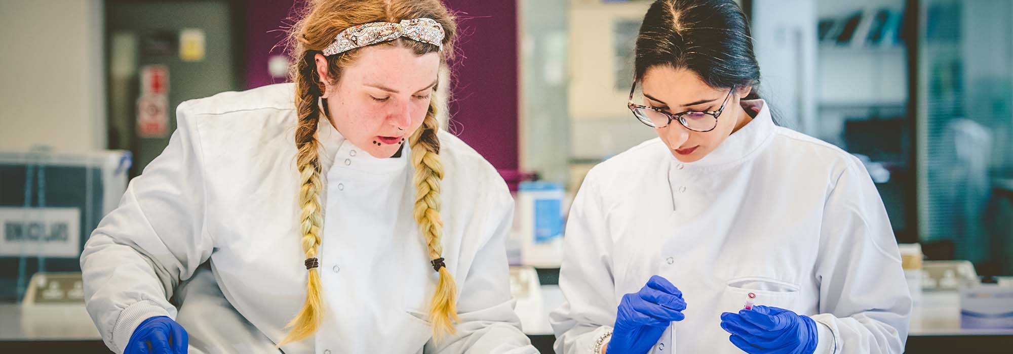 two female students working in the lab, they are wearing white lab coats and surgical gloves.