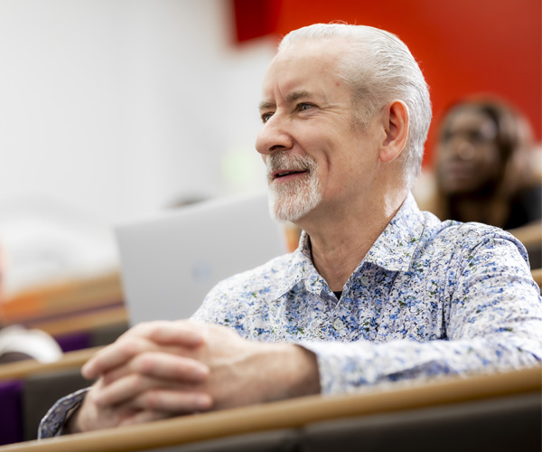 A mature man with a beard wearing a blue shirt is sitting in a lecture theatre