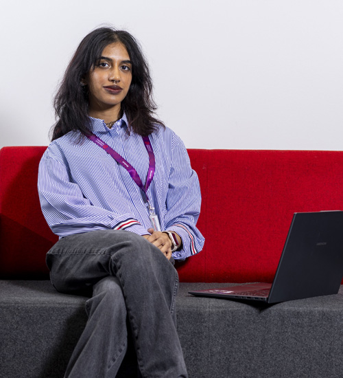 A young female student with dark hair, wearing a blue shirt, is sitting confidently with her laptop