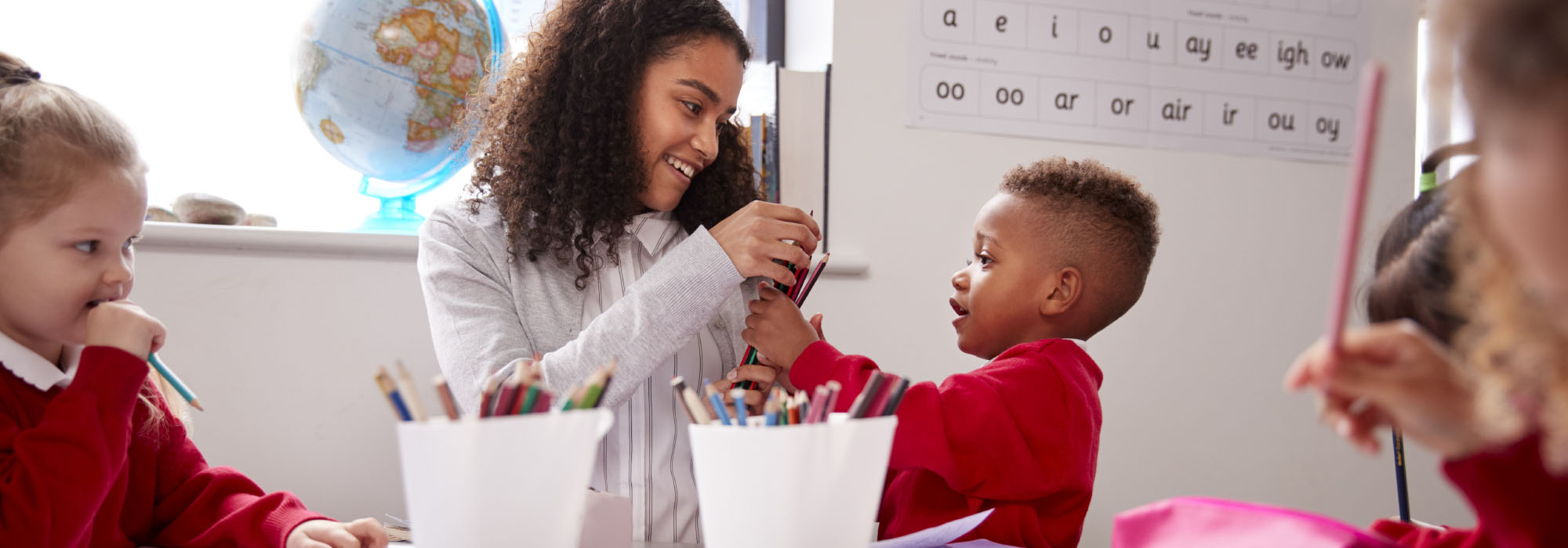 female trainee teacher helps a young student choose pencils in a classroom setting