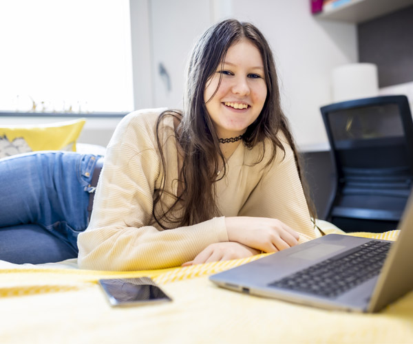 A young female student with long brown hair is casually using her laptop in her student accommodation room
