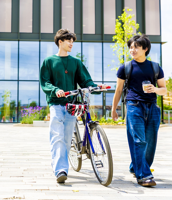 Male students are walking outside the student hub, one is pushing a bicycle