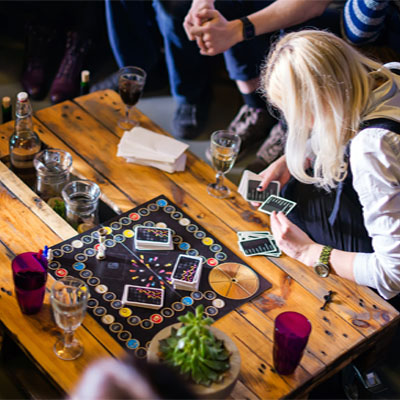 A group of people playing a table top card game on a wooden table with a selection of beverages