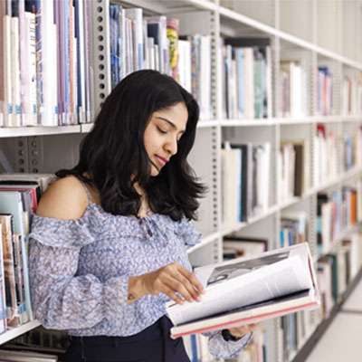 A female wearing a blue top is reading a book while standing next to the book shelf in the university library