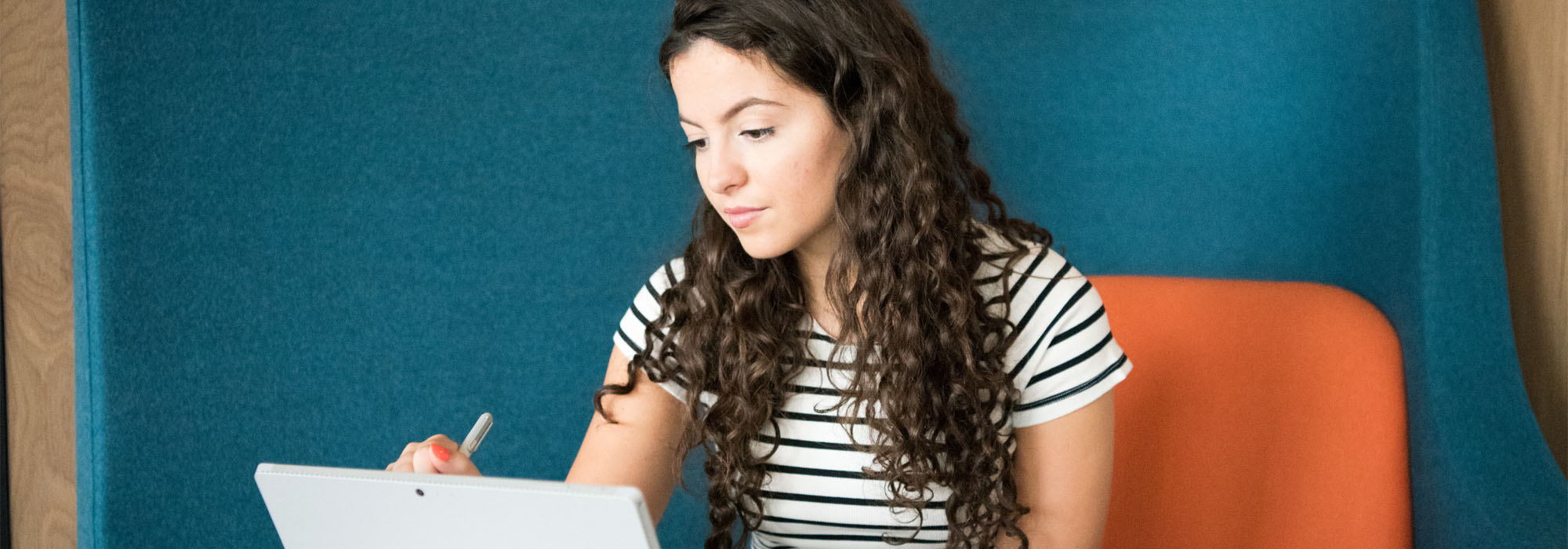 A female wearing a white and black striped t-shirt sitting on a blue chair studying on a laptop